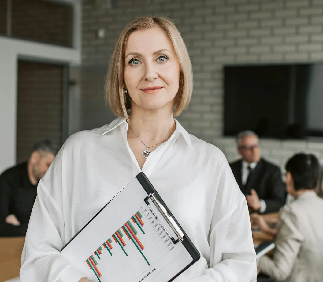 Smiling woman in white shirt holding a clipboard with colorful bar charts, with colleagues discussing in background.